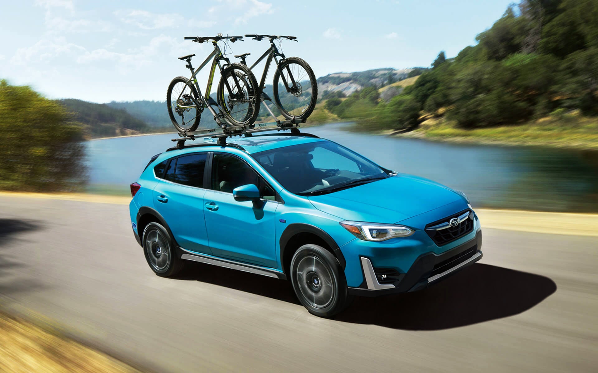 A blue Crosstrek Hybrid with two bicycles on its roof rack driving beside a river | Dalton Subaru in National City CA