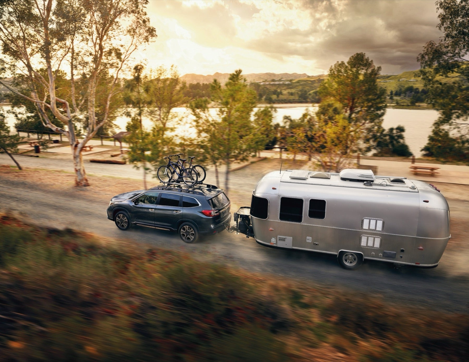 Gray Subaru Ascent with roof-mounted bikes towing a silver Airstream trailer past a lake at golden hour, the kind of Subaru compared across Premium and Limited trims at Dalton Subaru National City
