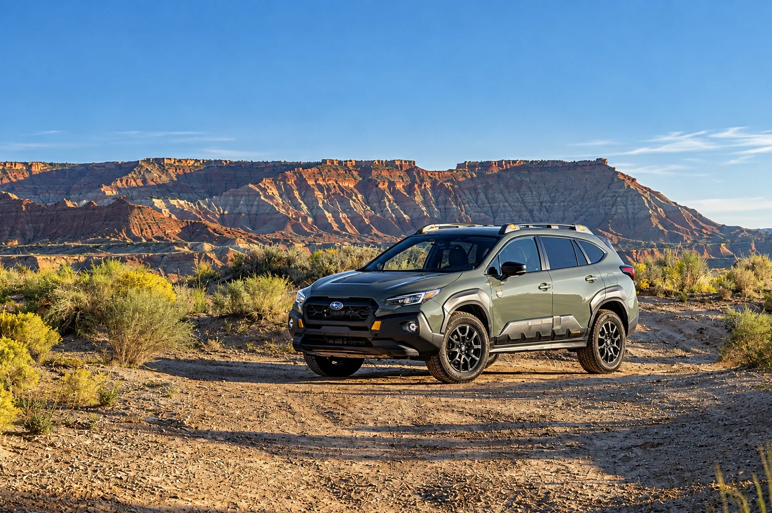 A green Subaru Wilderness model parked on a desert trail below red mesas.