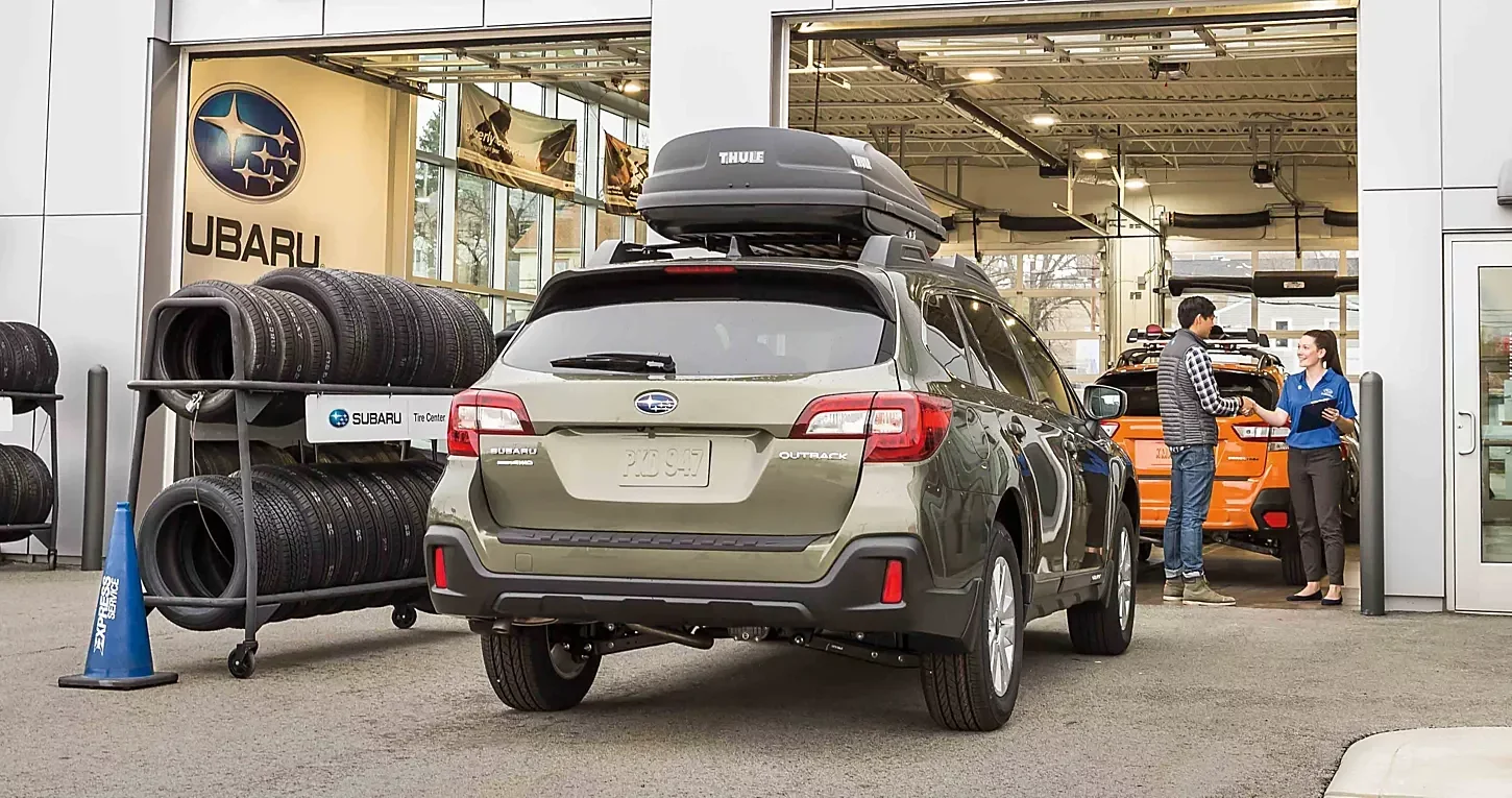 A Subaru Outback with a roof box in the service drive, beside a tire rack.