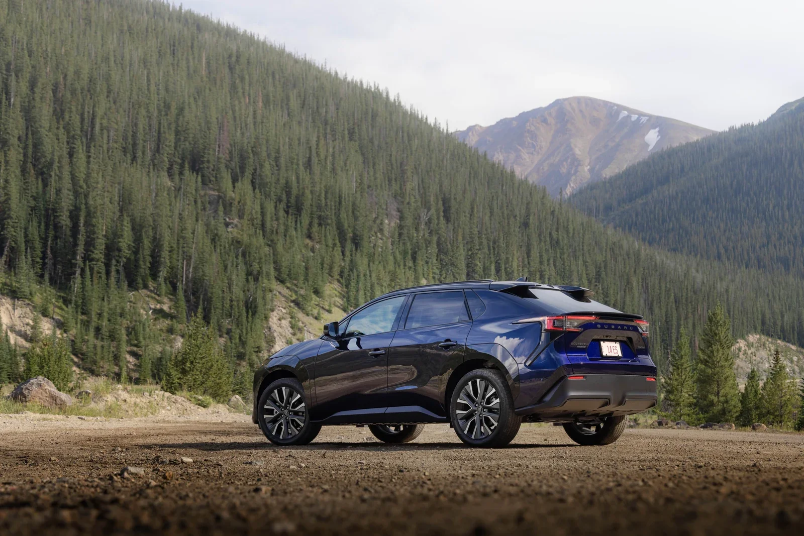A dark blue Subaru Solterra parked on gravel below forested mountains.