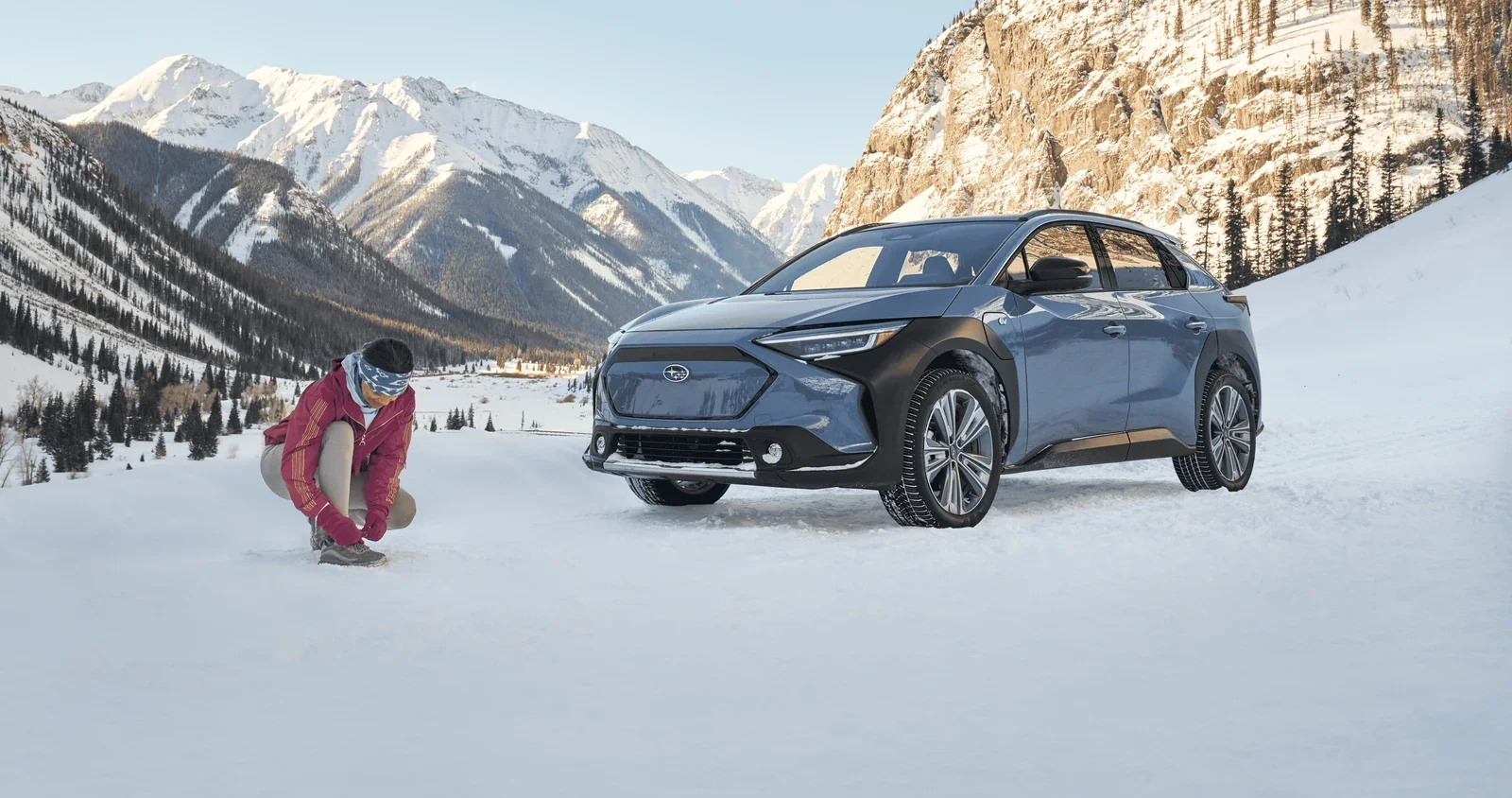 A Subaru Solterra parked in snow below the mountains, with a woman kneeling beside it.