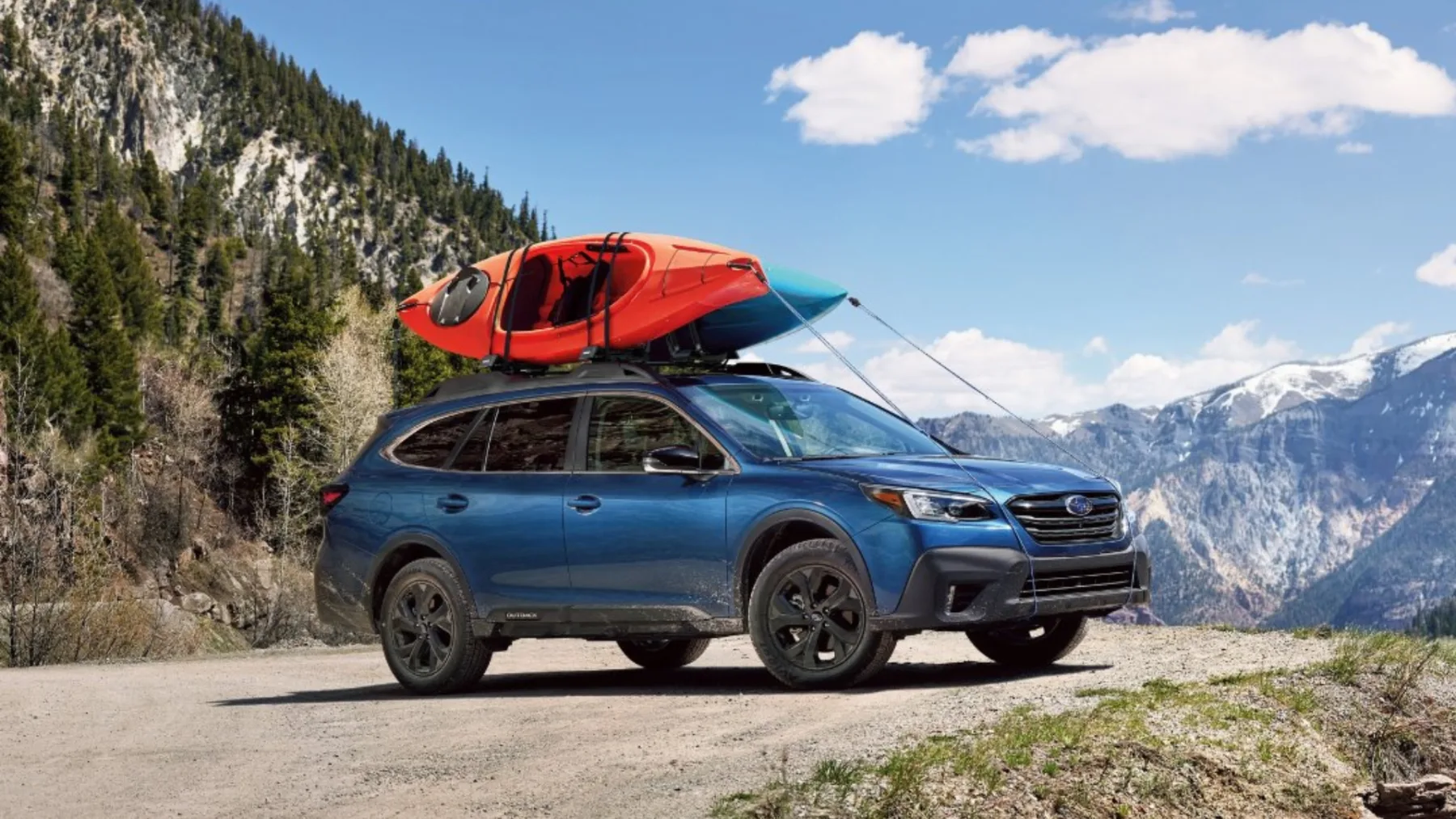 A blue Subaru Outback with kayaks on the roof rails parked on a mountain road, the kind of use Outback buyers lease or finance one for