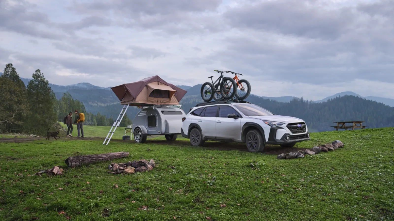 A white Subaru Outback with mountain bikes on the roof rack, towing a teardrop trailer at a mountain campsite